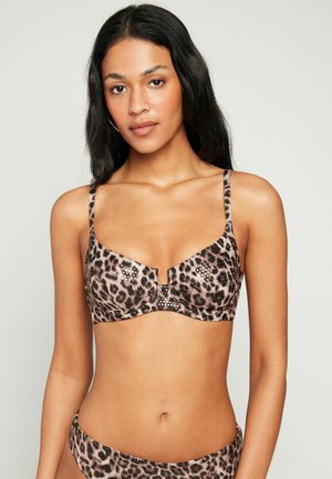 Frau mit langen schwarzen Haaren, die einen Leopardenmuster-BH und passende Unterteile vor einem schlichten hellen Hintergrund trägt.