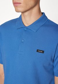 Polo bleu en coton avec col côtelé, patte à deux boutons, et une petite étiquette logo Calvin Klein noire sur la poitrine.