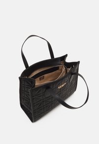 Sac fourre-tout matelassé noir avec deux anses, ouverture supérieure montrant la doublure intérieure beige et une poche zippée, logo Guess doré sur le devant.