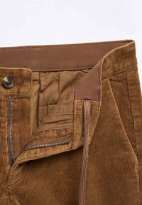 Pantalon en velours côtelé marron avec fermeture éclair partiellement ouverte, bouton fermé et doublure intérieure visible.