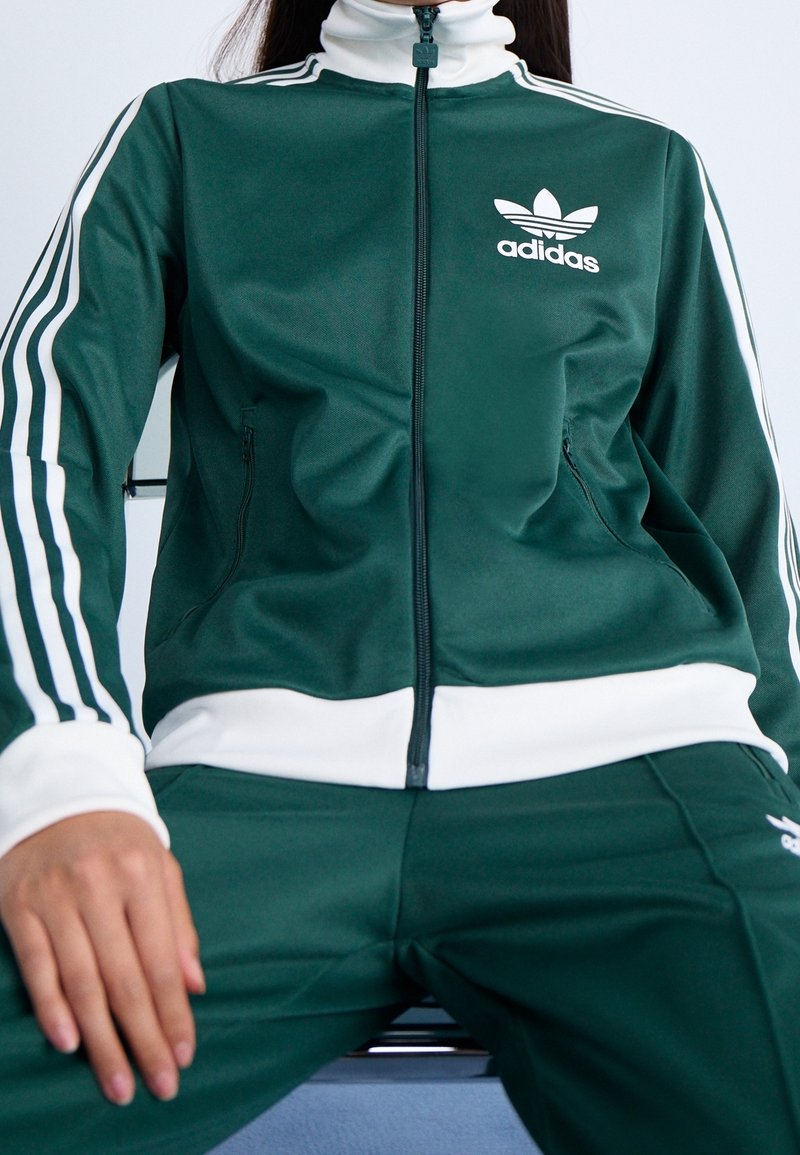 Person som bär en mörkgrön Adidas träningsdräkt med vita ränder och logo, sittande med händerna vilande på låren mot en ljus bakgrund.