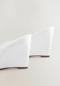 Sandales à talons compensés blanches avec une surface lisse, un design incurvé et un talon surélevé, présentant un motif découpé minimaliste sur les brides.