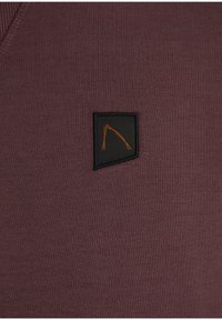 Maglione a scollo a V in bordeaux intenso con texture liscia. Presenta una piccola toppa di tessuto nera con un disegno geometrico arancione.