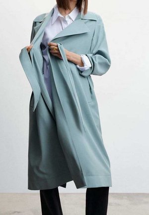 Vrouw die een lichtblauwe lange trenchcoat aanpast die gedragen wordt over een wit overhemd en zwarte broek, tegen een effen achtergrond.