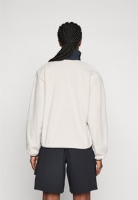 Person med flettet hår i lav hestehale, iført en hvit fleecejakke og svarte shorts, som står med ryggen til mot en ensfarget bakgrunn.