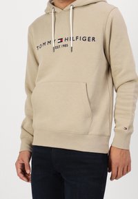 Beige hoodie with a front kangaroo pocket, drawstrings, and a logo text "TOMMY HILFIGER" with an embroidered flag on the cuff.