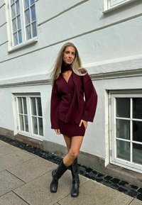 Burgundská pletená kardigan s širokými rukávy, vrstvená přes odpovídající pletené šaty, doplněná černými kozačkami po kolena, na pozadí bílé zdi.