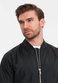 Veste bomber noire avec une texture lisse et brillante. Équipée d'une fermeture éclair, d'un col côtelé et de poignets élastiques. Design simple et minimaliste.