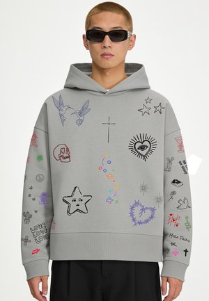 Personne portant un sweat à capuche gris avec divers gribouillis colorés, comprenant des étoiles, des cœurs, des oiseaux, un crâne et des symboles, assorti à des lunettes de soleil noires.
