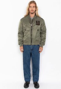 Olivgrüne Bomberjacke mit Reißverschluss vorne, gerippten Bündchen und Saum. Verfügt über einen Aufnäher auf der oberen linken Brust und zwei Seitentaschen.