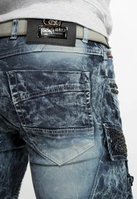 Jean en denim bleu délavé avec plusieurs poches, des rivets décoratifs et une étiquette logo. Texture usée et détails de couture légers visibles.