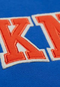 Blauem Stoff mit großen roten und weißen gestickten Buchstaben "K" und "N." Die Textur ist weich und hat eine glatte Oberfläche mit kontrastierenden Farben.