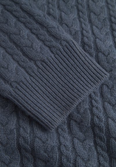 Detail tmavě modrého pleteného svetru s kabelovým vzorem a žebrovaným lemem, který ukazuje jemnou texturu a vrstvené designové prvky.