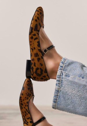 Bailarinas de estampado de leopardo con una correa negra brillante en el tobillo y punta cuadrada. El material tiene una textura suave, complementada por un bajo tacón cuadrado.