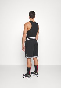Homme debout de dos portant un haut noir sans manches, un short de basketball noir avec des rayures blanches, des chaussettes noires avec des accents rouges, et des baskets.