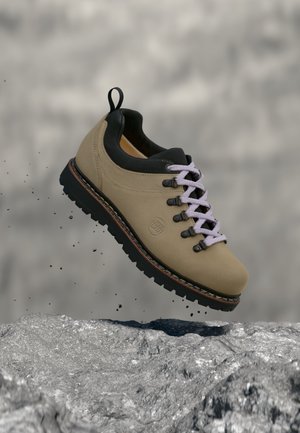 Beiger Wanderschuh mit schwarzer Sohle und weißen Schnürsenkeln über einer felsigen Oberfläche, kleine Partikel schweben herum, als ob er im Sprung wäre.