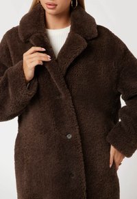 Abrigo de borreguillo marrón con cuello de muesca y grandes botones, que presenta una tela suave y texturada, además de un corte holgado.