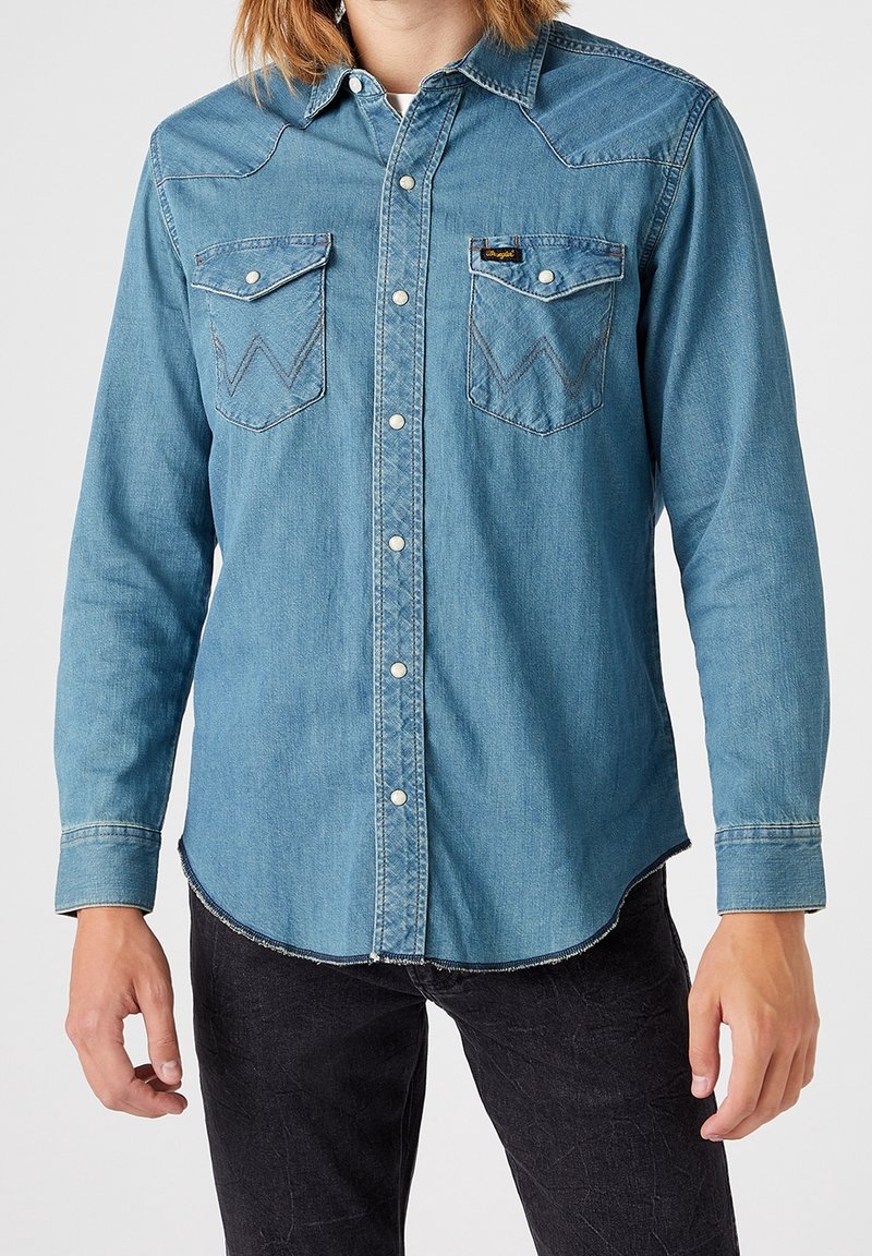 Homme portant une chemise en denim bleu clair à boutons avec deux poches sur le devant, sur un pantalon noir, debout devant un fond uni.