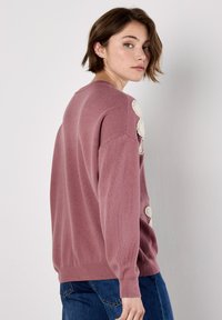 Rosa gestrickter Pullover mit einem lockeren Schnitt, der dekorative cremefarbene Häkelakzente an den Schultern sowie gerippte Bündchen und Saum aufweist.