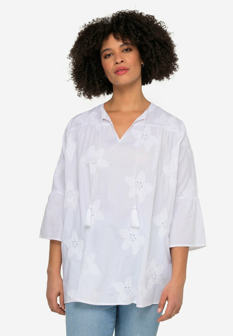 Hvid bomuldsbluse med 3/4 klokkeærmer, der har blomsterbroderi og frynser ved halsudskæringen. Løs pasform med V-hals design.