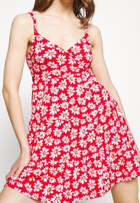 Czerwona sukienka bez rękawów z dopasowanym korpusem i rozkloszowanym dołem, ozdobiona białym wzorem stokrotek z żółtymi środkami na jednolitym tle.