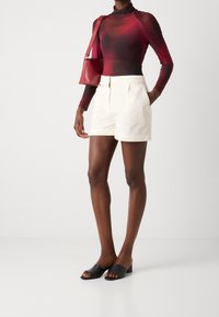 Modelka na sobě má burgundský průhledný top s dlouhým rukávem, bílé přizpůsobené šortky, černé sandály na otevřenou špičku na podpatku a nese červenou kabelku přes rameno.