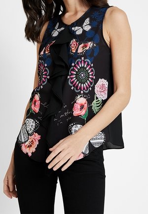 Mouwloze zwarte blouse met kleurrijke bloemen- en vlinderpatronen en een gerimpelde voorkant, gedragen met zwarte broek.