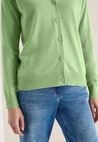 Personne portant un cardigan boutonné vert clair et un jean bleu, debout les mains détendues le long du corps.