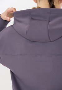 Person som bär en lila hoodie, justerar huvan, med en enda fläta synlig på baksidan av huvudet mot en enfärgad bakgrund.