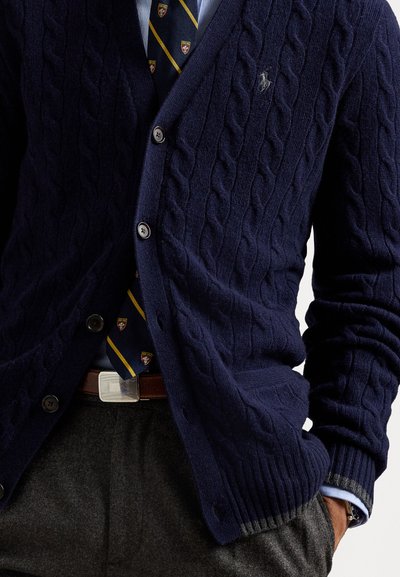 Cardigan en tricot marin avec un col en V, six boutons et des poignets côtelés, porté sur une chemise bleu clair et une cravate à motifs. Pantalon gris et ceinture visibles.