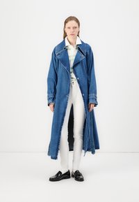 Dolga jeans jakna v bogatem modrem odtenku z velikim ovratnikom in odprtim prednjim delom. Nosi se čez črtasto srajco in bele hlače, z črnimi mokasini.