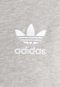 Pilkas audinys su siuvinėtu baltu Adidas logotipu ir klasikiniais trimis juostelėmis virš prekės pavadinimo. Švelni tekstūra ir glotnus paviršius.