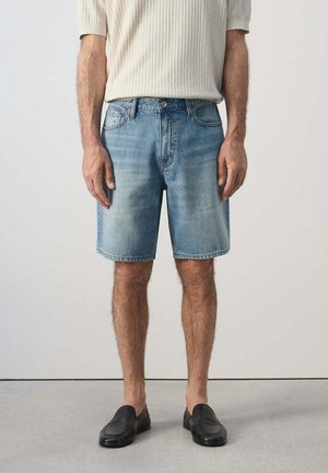 Uomo che indossa pantaloncini di denim azzurro chiaro, maglietta crema a maniche corte a coste e scarpe nere slip-on, in piedi davanti a uno sfondo semplice.