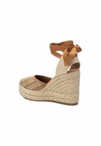 Zapato de cuña de yute en tono arena con parte superior tejida y texturizada, parte trasera abierta y una gruesa plataforma de yute. Cuenta con un lazo en el tobillo de color marrón.