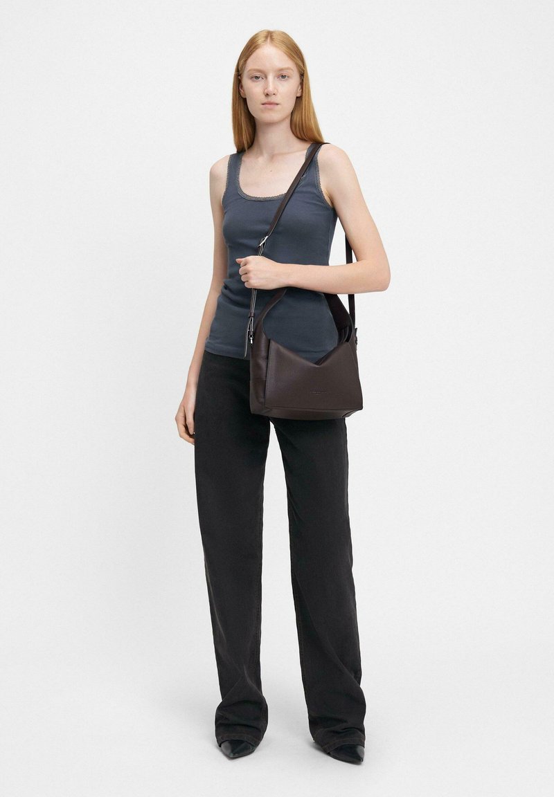 Borsa a mano in pelle marrone con una forma strutturata, tracolla regolabile e una texture liscia. Indossata con una canotta grigia e pantaloni larghi neri.