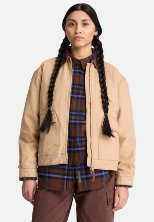 Junge Frau mit langen Zöpfen, die eine beige Jacke über einem braun-blau karierten Hemd und braune Hosen trägt, steht vor einem weißen Hintergrund.