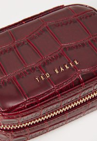 Bolso de cuero de charol rojo oscuro con textura de cocodrilo, cierre dorado y "TED BAKER" en relieve en letras doradas en la parte superior.