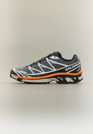 Schwarzer Sportschuh mit strukturierter Oberfläche, weißen Akzenten, orangefarbener Zwischensohle und gemustertem Laufsohlenprofil. Die Schnürsenkel sind deutlich sichtbar.