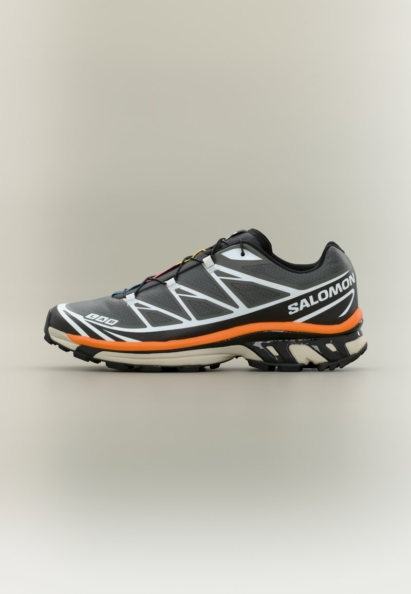 Μαύρο αθλητικό παπούτσι με υφή, με λευκές λεπτομέρειες, πορτοκαλί ενδιάμεση σόλα και σχεδιασμένη σόλα. Οι κορδόνια είναι εμφανή.