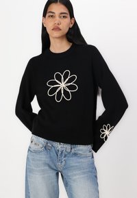 Svart strikkegenser med brodert kremfarget blomstermønster på fronten og ermene. Har rund hals og ribbestrikket kant. Brukes sammen med lyseblå jeans.