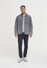 Denim jakna u izblijedeloj sivoj boji s kopčanjem na gumbe i jednim džepom na prsima; uparena s tamnim hlačama i svijetlim tenisicama na jednobojnoj pozadini.