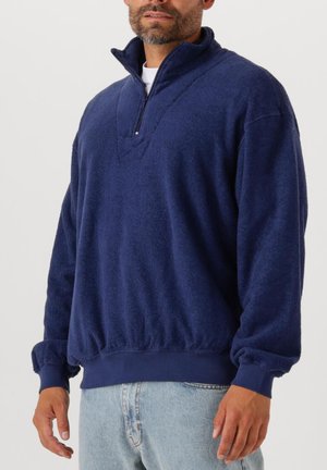 Mann trägt einen marineblauen Fleecepullover mit Viertelreißverschluss und hellblaue Jeans, steht vor einem schlichten hellen Hintergrund.