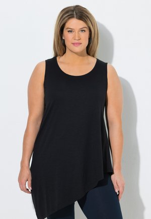Femme aux cheveux châtain clair portant un haut noir asymétrique sans manches et un pantalon foncé, debout devant un mur blanc uni.