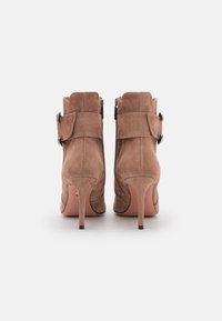 Bottines en daim marron clair avec un talon haut effilé, présentant des zips sur les côtés et une large sangle décorative avec une boucle en métal.