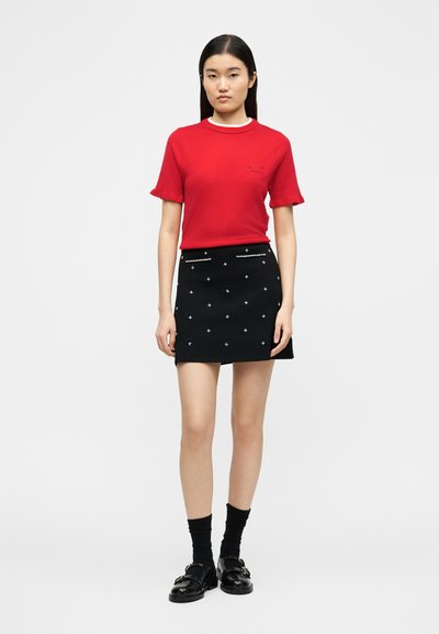 Γυναίκα που στέκεται φορώντας κόκκινο τοπ με κοντά μανίκια, μαύρη μίνι φούστα με λευκές διακοσμήσεις, μαύρες κάλτσες και μαύρα μοκασίνια σε λευκό φόντο.