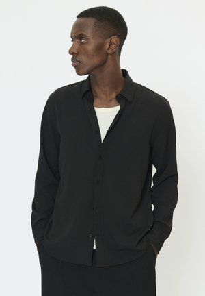 Sort skjorte med knapper, lavet af tekstureret stof, lange ærmer og afslappet pasform. Båret over en hvid T-shirt med rund hals.