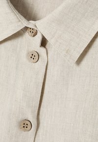 Camicia in lino beige con colletto a punta e cinque bottoni in legno naturale con quattro fori ciascuno, che mette in mostra una superficie di tessuto testurizzato.