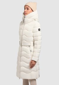 Geacă puffer albă cu un design ondulat, guler înalt și talie cu curea. Prezintă un logo pe mânecă și buzunare cu fermoar.