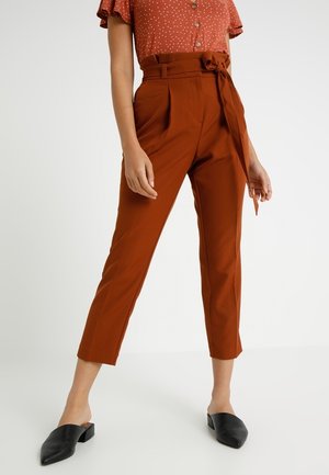 Vrouw draagt roestkleurige hooggetailleerde taps toelopende broek met een tailleband, gecombineerd met een roestkleurig overhemd met witte stippen en zwarte instapschoenen.