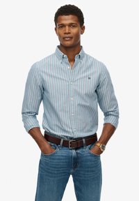 Overhemd met blauwe en witte verticale strepen; kraag, lange mouwen, subtiel logo; gecombineerd met blauwe denim en een bruine riem.